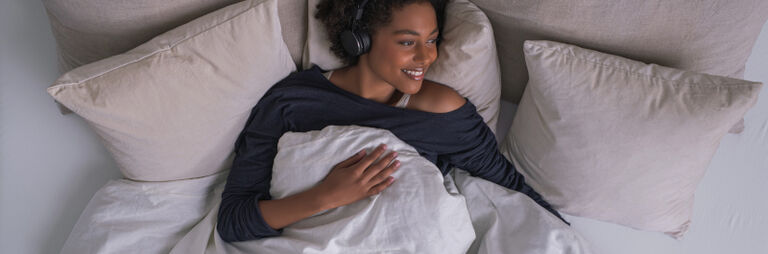 Vrouw ligt stralend op bed naar muziek te luisteren met een koptelefoon op.